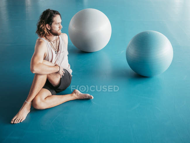 D'en haut de l'homme barbu en vêtements de sport étirant les jambes croisées sur le sol bleu avec des balles de gymnastique en studio — Photo de stock