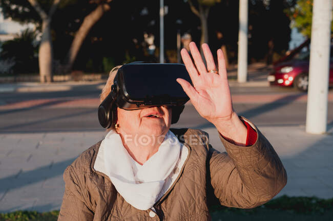 Nonna lungimirante in cuffia realtà virtuale vedere nuovo mondo simulato. Donna anziana toccare oggetto inesistente con mano sollevata in piedi su sfondo sfocato con la strada della città — Foto stock