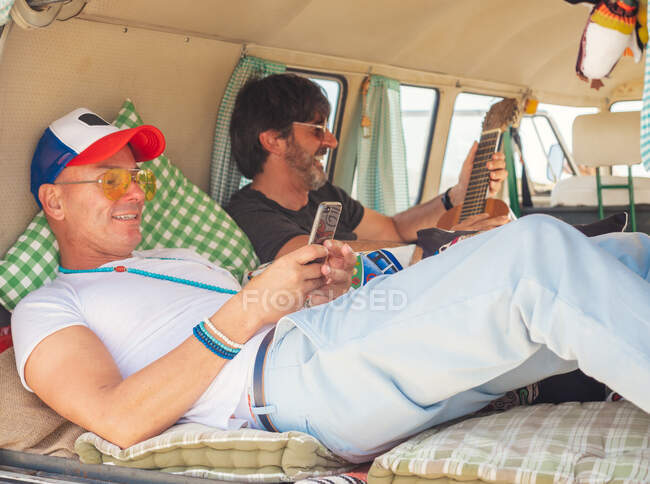 Вид сбоку улыбающихся мужчин, разглядывающих гаджеты в симпатичном уютном трейлере — стоковое фото