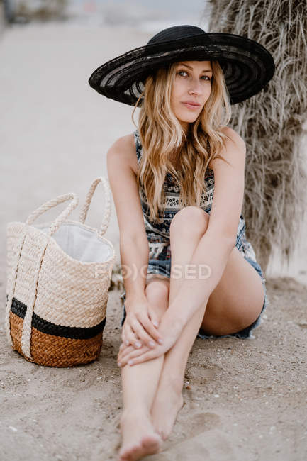 Blondine mit schwarzem Hut sitzt mit Sommertasche auf Sand und blickt in die Kamera — Stockfoto