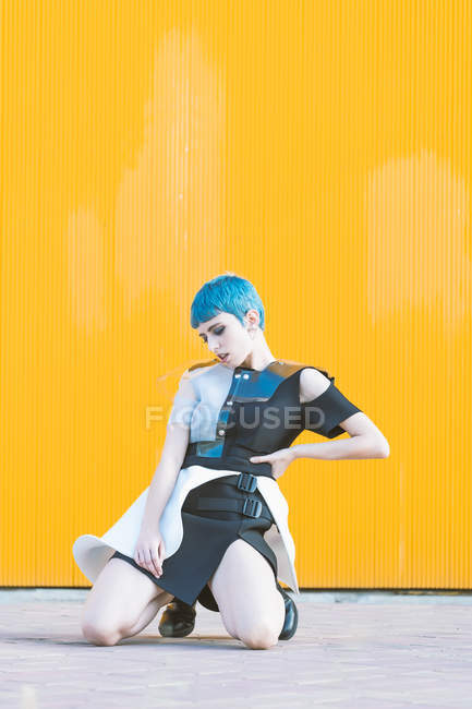 Mujer joven en vestido futurista de moda mirando hacia abajo mientras está sentado en las rodillas en el pavimento contra la pared de color amarillo brillante - foto de stock