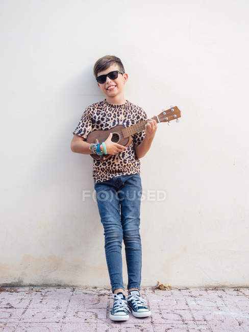 Милый радостный энергичный веселый ребенок в джинсе и праздничной футболке, веселый играя на укулеле на фоне белой стены — стоковое фото