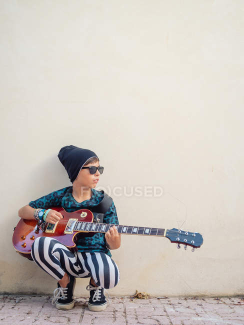Frech aktiver aufgeregter Junge in bunten Klamotten, der Gitarre spielt, angelehnt an den Hintergrund einer weißen Wand — Stockfoto