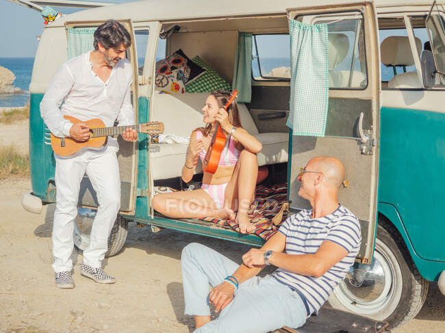 Fröhlich entspannte Menschen genießen das Spielen von Ukulele und Gitarre in der Nähe von Wohnwagen am staubigen Meer vor dem Hintergrund des erstaunlichen Wassers bei hellem Tag — Stockfoto