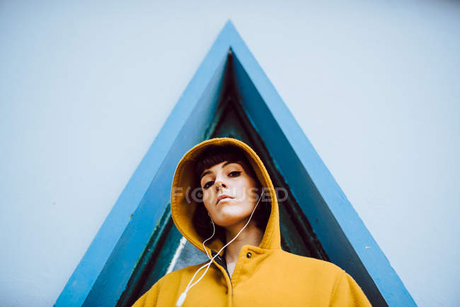 Молодая женщина в желтом теплом пальто слушает музыку и смотрит в камеру, стоя напротив треугольного окна и серой стены здания — стоковое фото