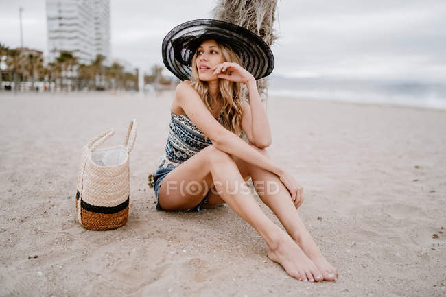 Mulher loira de chapéu preto sentado na areia com saco de verão e olhando para longe pensativo — Fotografia de Stock
