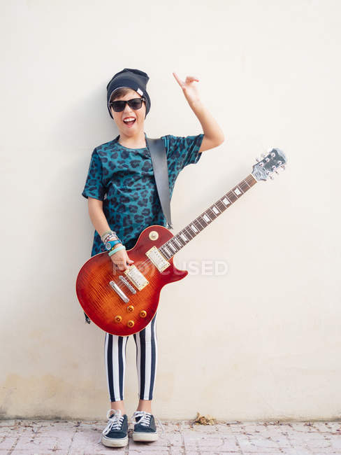 Cheeky actif excité garçon en vêtements colorés jouer de la guitare, montrant deux doigts vers le haut sur le fond de mur blanc — Photo de stock