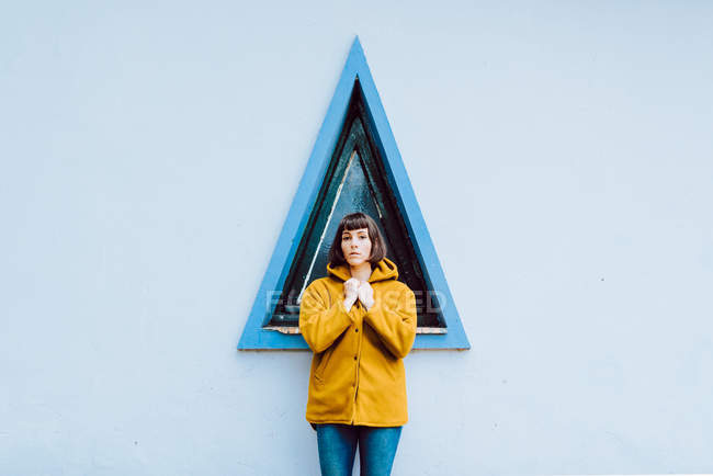 Молодая женщина в желтом теплом пальто улыбается и смотрит в камеру, стоя напротив треугольного окна и серой стены здания — стоковое фото