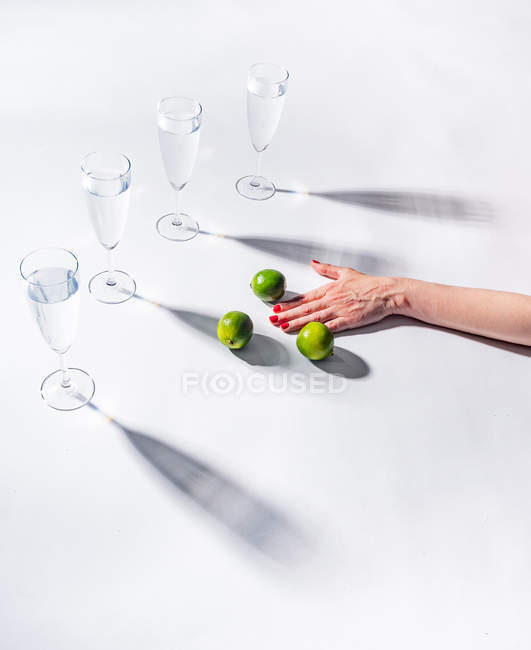 Жіноча рука з зеленими стиглі Лайми біля чітких келихів води на білому фоні — стокове фото