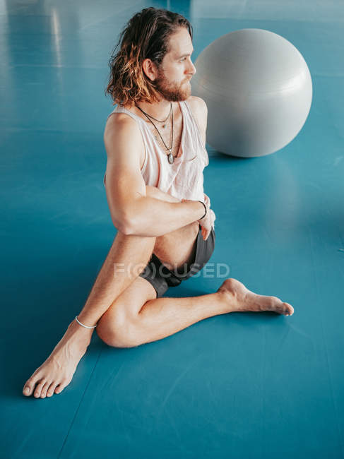 Зверху бородатого чоловіка в спортивному одязі, що розтягується ногами, схрещеними на синій підлозі з гімнастичними кульками в студії — стокове фото