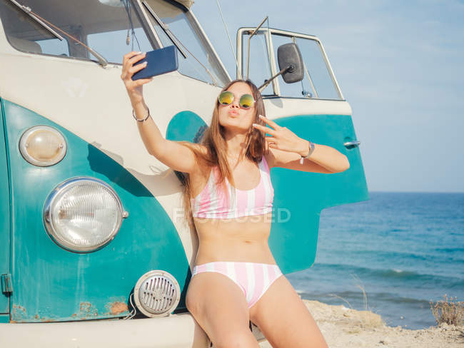 Atractiva mujer bronceada encantadora tomando selfie y sonriendo cerca de coche en la playa de arena en el día brillante - foto de stock