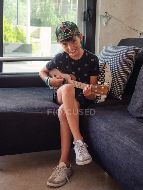 Sorrindo menino criativo jogando ukulele, sentado em sofá confortável e olhando para a câmera — Fotografia de Stock