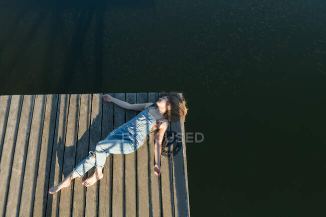 Vista superior da mulher bonita em maiô preto e chapéu deitado no cais de madeira do lago no céu azul claro e fundo da floresta — Fotografia de Stock