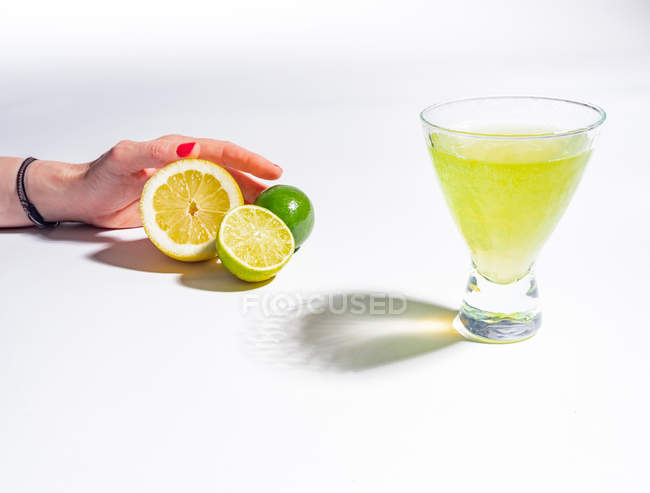 Female hand holding halved lemon and limes near glass of yellow lemonade drink on white background — Stock Photo