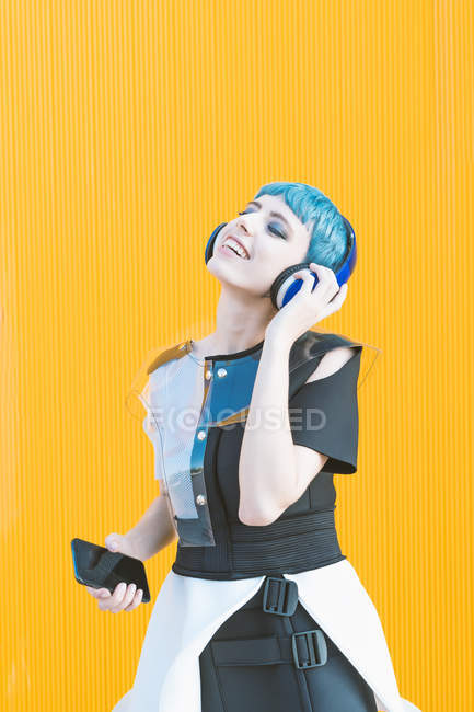 Mujer informal alegre en auriculares y teléfonos inteligentes escuchando música mientras está de pie contra la pared amarilla vívida - foto de stock