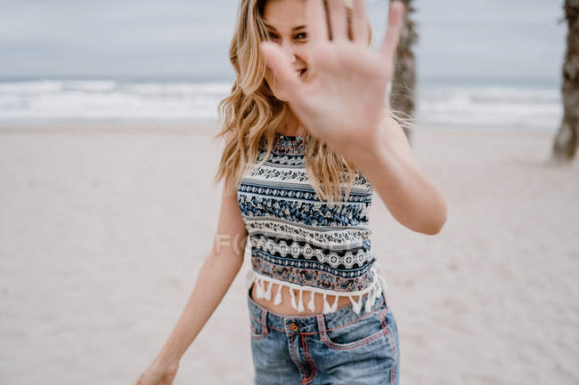 Femme blonde joyeuse en haut coloré et short en jean bloquant son visage avec la main sur le bord de la mer — Photo de stock