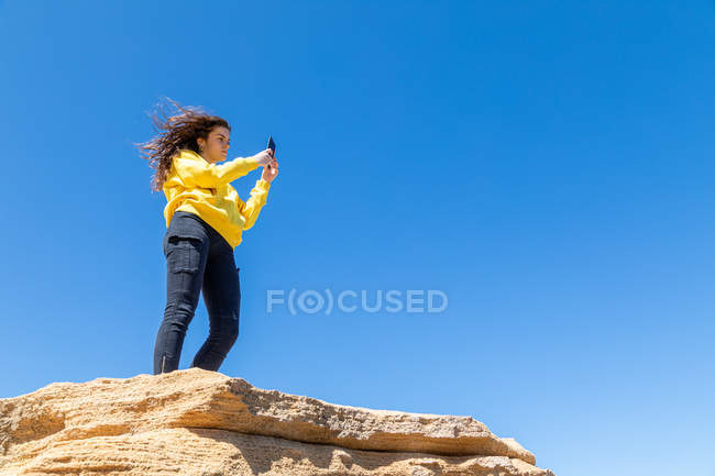 Молода брюнетка фотографує зі смартфоном, стоячи на скелі з пісковиком на фоні блакитного неба — стокове фото