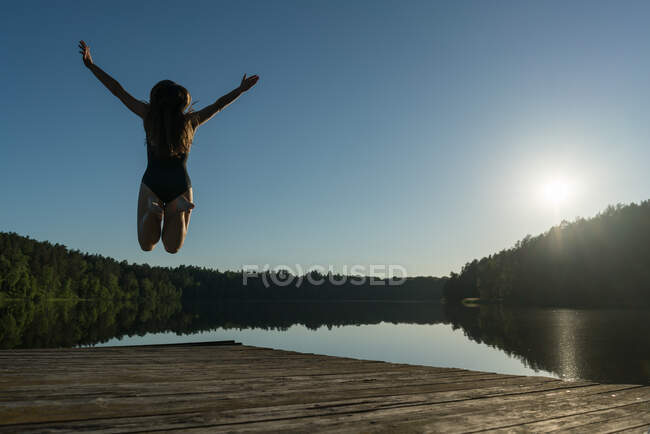 Back view of unrecognizable female in swimsuit jumping on wooden pier near forest lake raising hands up in sunny weather with clear blue sky — Stock Photo