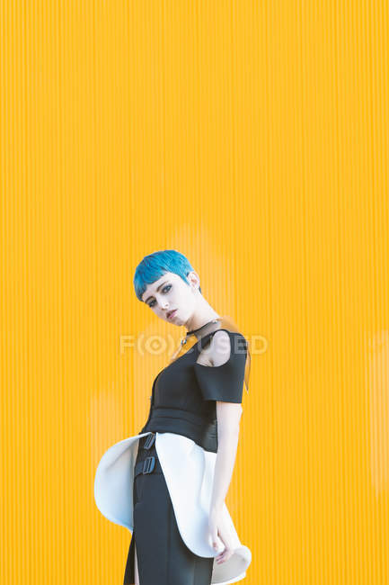 Young woman in trendy futuristic dress looking at camera while standing on knees on pavement against bright yellow wall — Stock Photo
