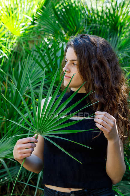 Portrait of young brunette woman in tropical bushes holding palm tree leaf — Stock Photo