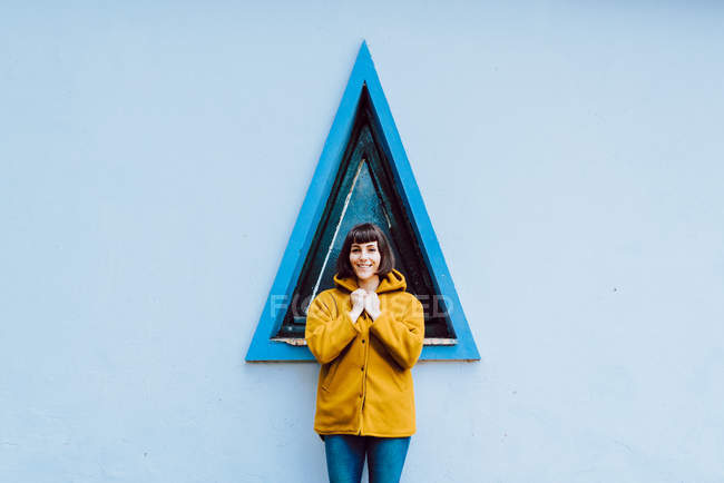 Mujer joven en abrigo cálido amarillo sonriendo y mirando a la cámara mientras está de pie contra la ventana del triángulo y la pared gris del edificio - foto de stock
