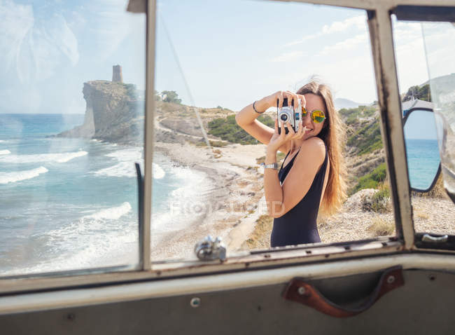 Young long haired woman with camera in swimsuit at sandy beach with foamy waves while looking at camera — Stock Photo