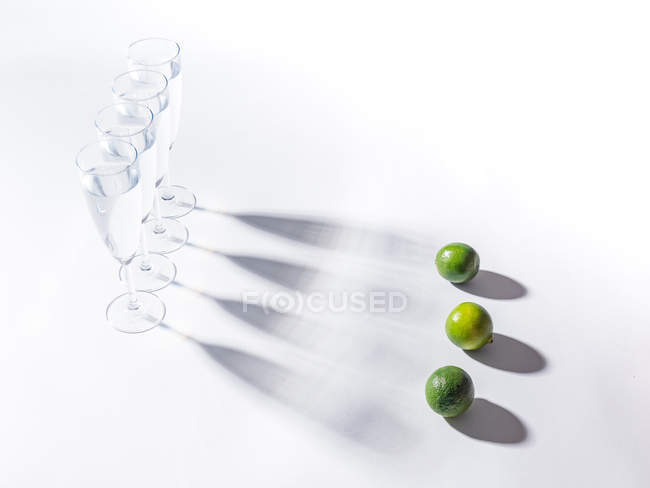 Sombras contrastantes de vasos de agua y limas verdes maduras sobre fondo blanco - foto de stock