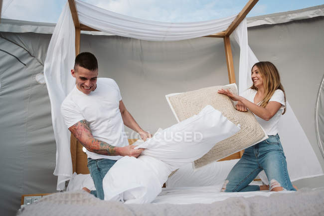 Jovem casal alegre se divertindo durante a luta travesseiro na cama em grande barraca com telhado transparente — Fotografia de Stock