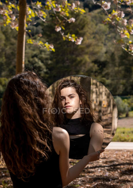 Back view of pretty brunette female in black top reflected in round mirror on nature background — Stock Photo