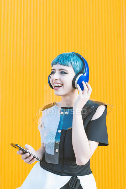 Mulher informal alegre em fones de ouvido e smartphone ouvindo música enquanto está de pé contra a parede amarela vívida — Fotografia de Stock