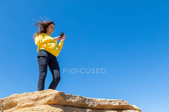 Молодая брюнетка женщина фотографируется со смартфоном, стоя на скале песчаника на фоне голубого неба — стоковое фото