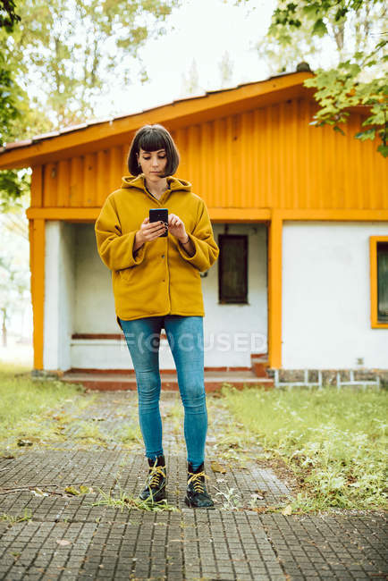 Junge Frau in lässigem Outfit lächelt und surft Smartphone, während sie an einem Herbsttag auf dem Land auf einem gefliesten Weg vor einem schönen Ferienhaus steht — Stockfoto