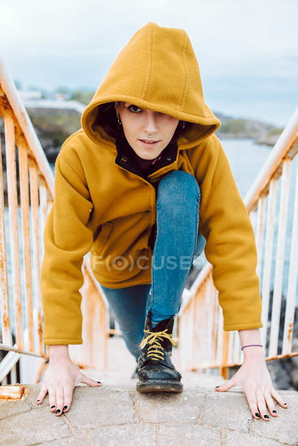 Jeune femme en veste jaune posant sur un escalier rouillé contre la baie avec de l'eau de mer ondulante — Photo de stock