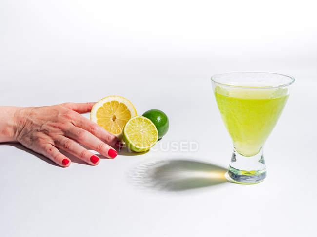 Mano femenina sosteniendo la mitad de limón y limas cerca del vaso de bebida de limonada amarilla sobre fondo blanco - foto de stock