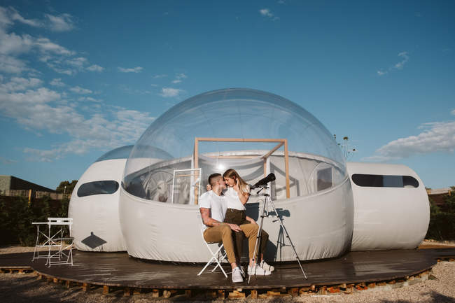 Femme élégante assise sur petit ami tour par télescope au ciel près de l'hôtel bulle — Photo de stock
