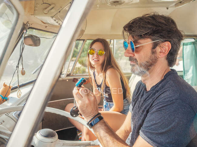 Hombre barbudo y mujer de pelo largo en gafas de sol de colores sentados en frente del mar del coche y mirando azul pequeño coche de juguete - foto de stock