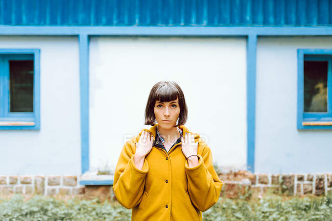 Mujer joven en abrigo cálido amarillo sonriendo y mirando a la cámara mientras está de pie contra el edificio de la pared gris - foto de stock