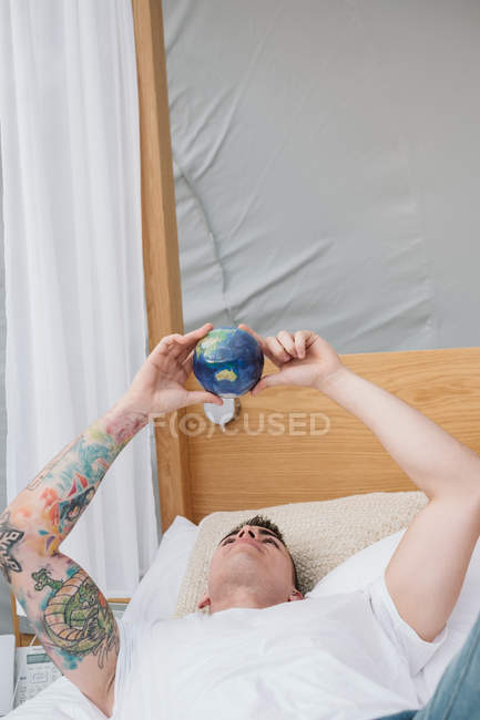 Handsome tattooed man in white t-shirt twisting in hands small earth ball while relaxing on bed — Stock Photo