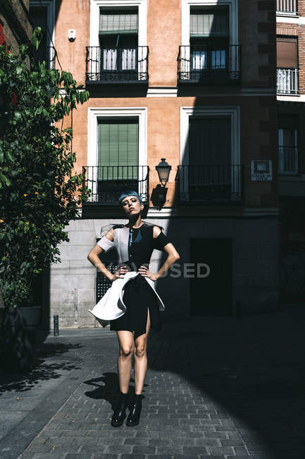 Mujer joven en vestido futurista de pie con las manos en la cintura en la calle contra el viejo edificio a la luz del sol - foto de stock