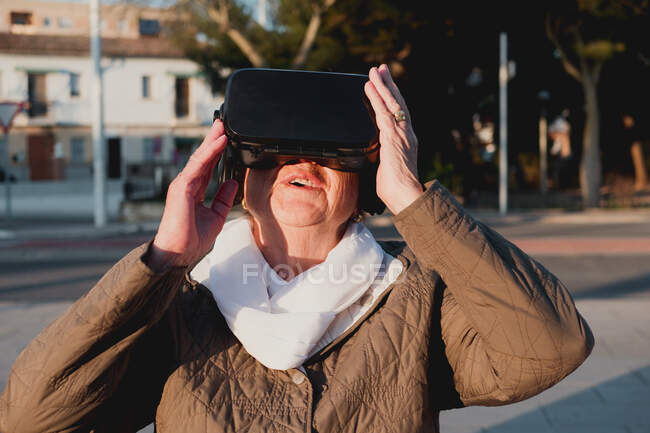 Nonna lungimirante in cuffia realtà virtuale vedere nuovo mondo simulato. Donna anziana toccare oggetto inesistente con mano sollevata in piedi su sfondo sfocato con la strada della città — Foto stock