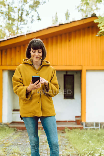 Jeune femme en tenue décontractée souriant et naviguant smartphone tout en se tenant sur le chemin carrelé à l'extérieur beau chalet le jour de l'automne dans la campagne — Photo de stock