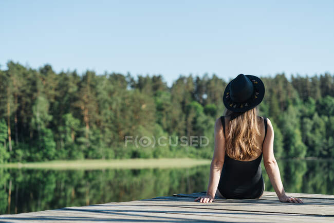 Visão traseira de mulher pensativa irreconhecível em maiô preto e chapéu sentado no cais de madeira e admirando vista do lago no céu azul claro e fundo da floresta — Fotografia de Stock