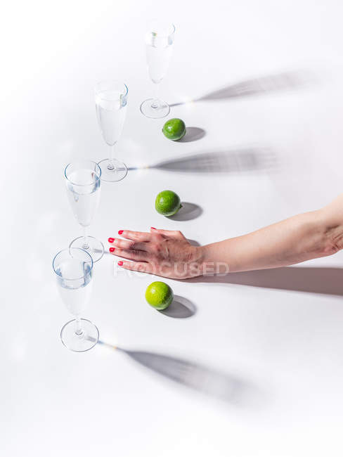 Mano femminile con lime verdi maturi vicino a vetri trasparenti d'acqua su sfondo bianco — Foto stock