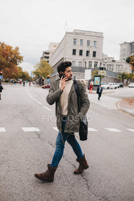 Trendy young man talking on mobile phone while walking on road on autumn day — Stock Photo