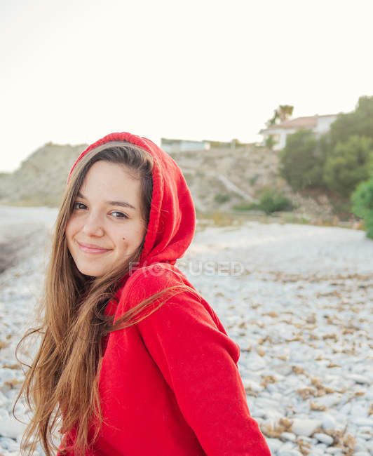 Charmante femme aux cheveux longs et au sweat à capuche rouge souriant et regardant la caméra sur le bord de la mer — Photo de stock