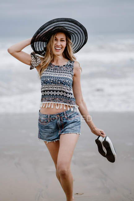Atractiva mujer en sombrero negro sosteniendo bolsa de playa y zapatos mientras disfruta de una vista pintoresca del océano mirando a la cámara - foto de stock