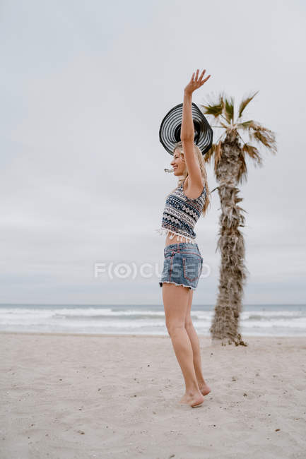 Привлекательная женщина в топе и шортах танцует на песчаном берегу с черной шляпой в поднятой руке — стоковое фото