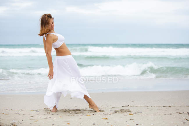 Anmutige junge Frau im weißen Outfit tanzt auf Sand in der Nähe des winkenden Meeres — Stockfoto