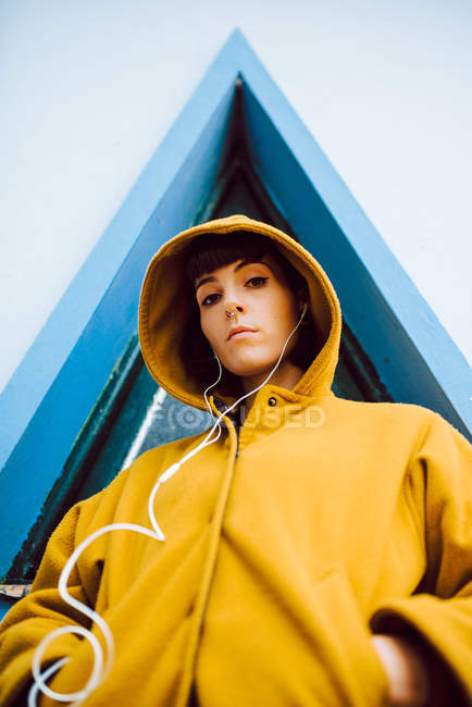 Young woman in yellow warm coat listening to the music and looking at camera while standing against triangle window and gray wall of building — Stock Photo