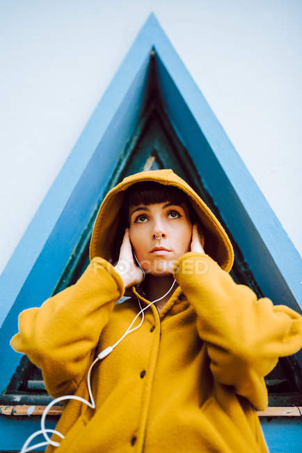 Young woman in yellow warm coat listening to the music and looking at camera while standing against triangle window and gray wall of building — Stock Photo